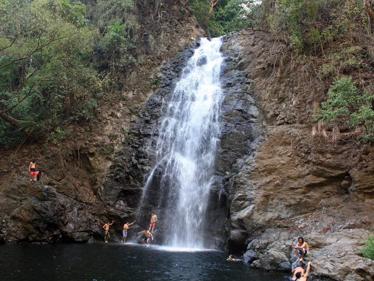 Swimmers in the pool at the base of the main Montezuma waterfall, with a tall white cascade dropping down dark rock